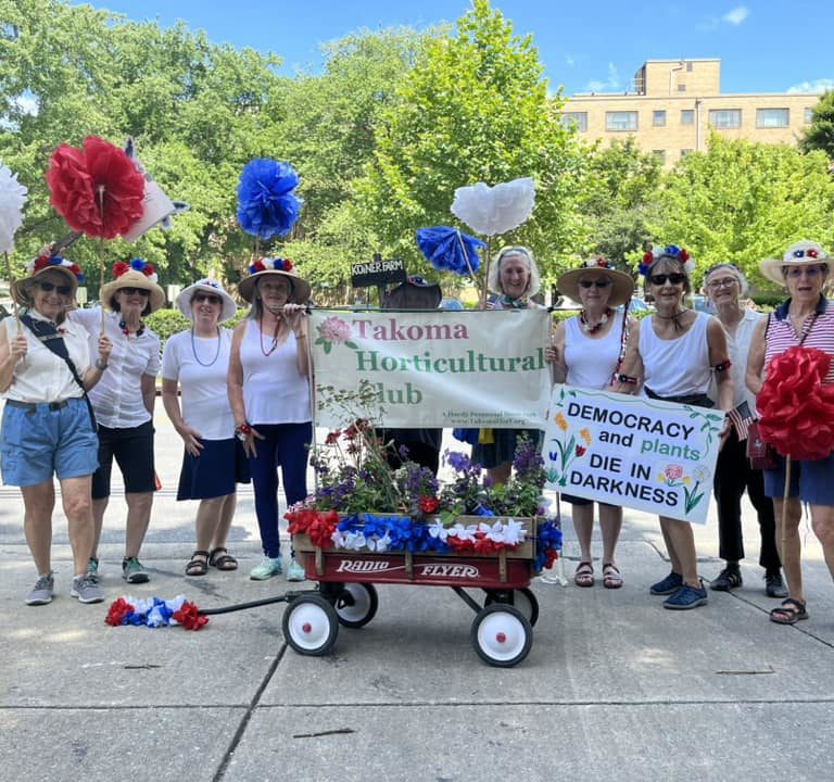 The THC marchers at the 2023 4th of July parade with a wagon full of red, white and blue flowers and a sign that says "Democracy and plants die in darkness".